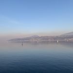 Our Instagram profile picture that is a landscape shot with deep blue water on bottom, a bright blue sky on the top with an orange glow in the middle. There is a strip of land with hills and trees in the middle.