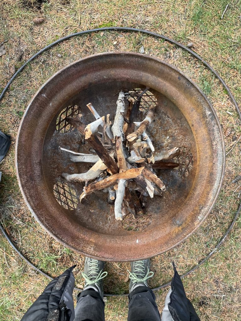 A pair of human legs and the end of a long jacket stand in front of a fire pit full of kindling ready to be lit. 
