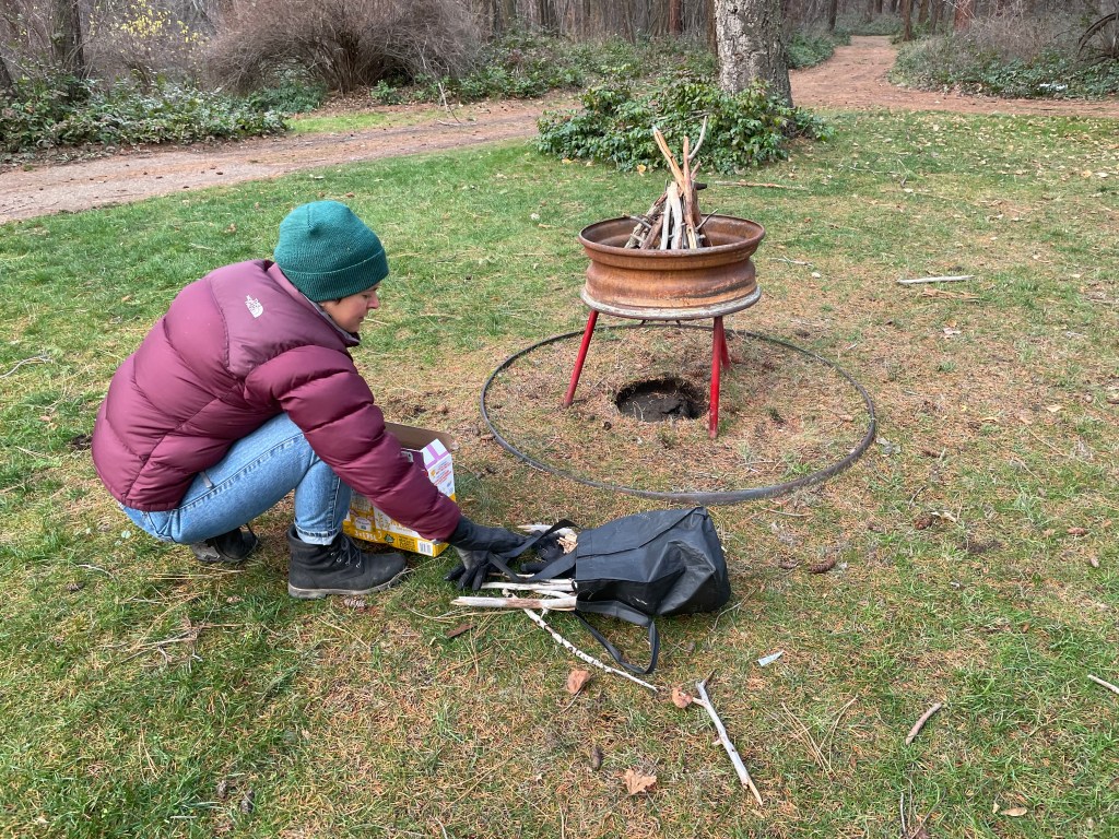 Human in in warm clothing crouches next to a pile of kindling and in front of a fire pit in which they are building a collection with which to start a fire. The pit is located in the middle of a metal ring on grass in a forest with a dirt road ringing the patch of grass. 