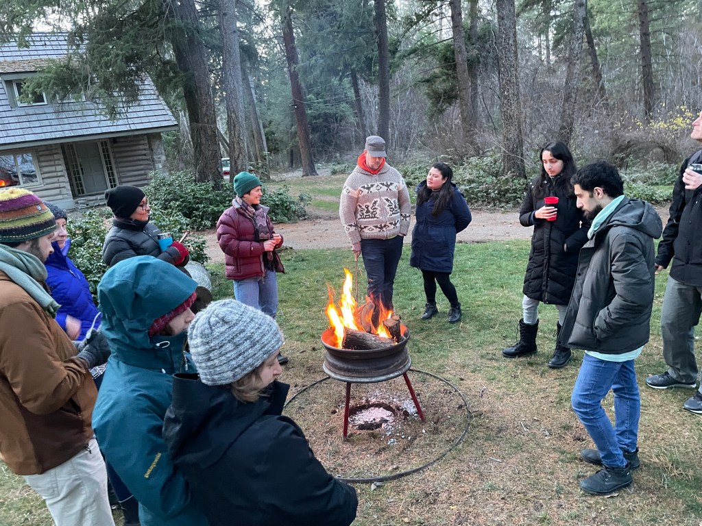 A group of humans dressed in warm clothing stand around a fire pit, lit, in a forest.