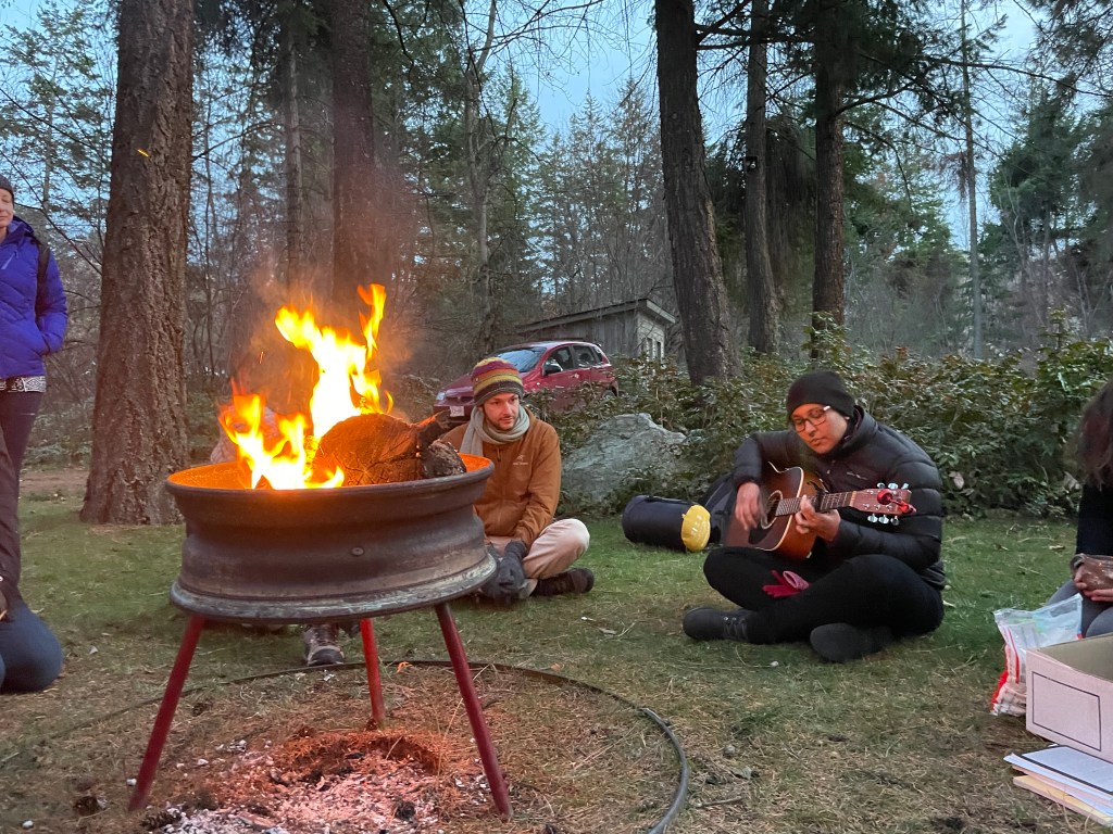 Two humans sit in front of a fire pit, one playing guitar, one listening. Others join from outside the frame. 