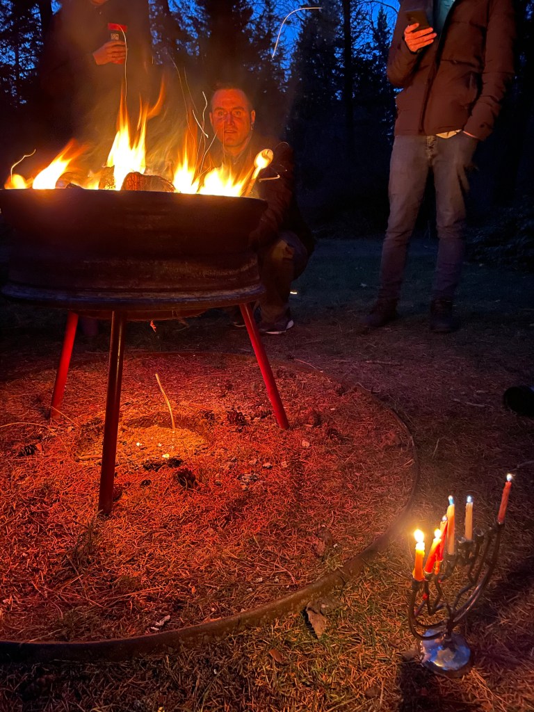 A manorah burns six candles next to the fire put, while one person's face is illuminated across the circle and another reads aloud from their phone.