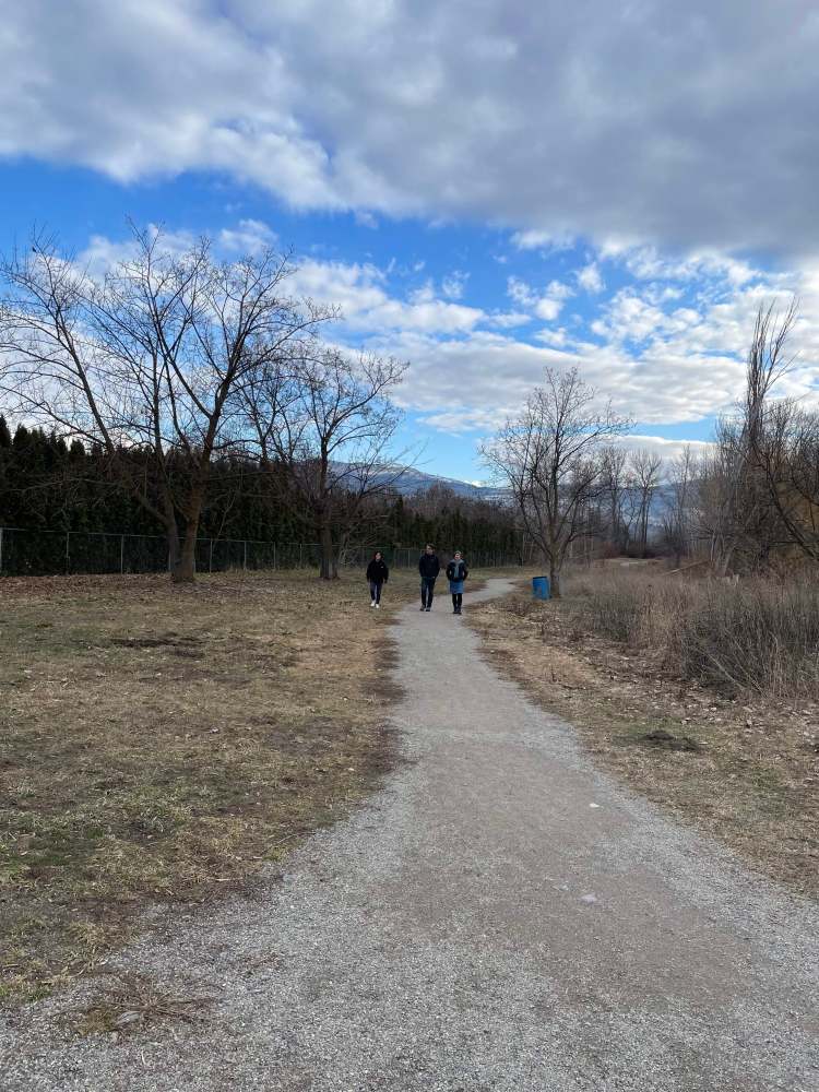 Three figures in the distance come towards us along a gravel path. Bare trees and dry grass are on either side of the grass, with mountains and a blue sky in the distance. 