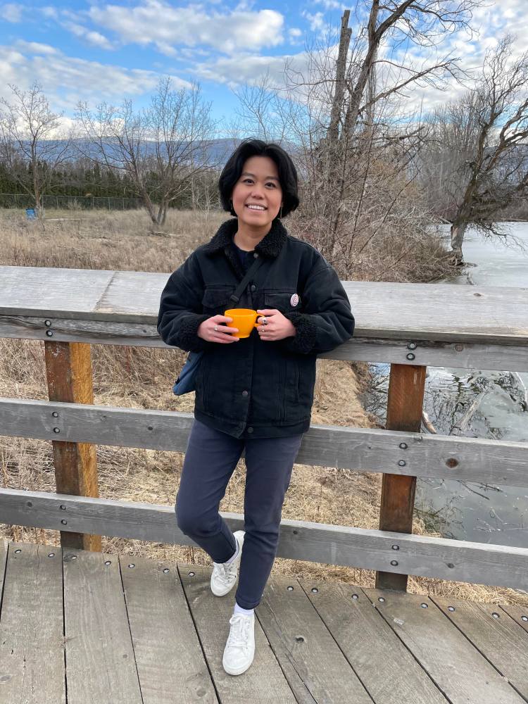 A smiling person with dark hair, a black coat and white running shoes leans against the wooden railing, and holds a bright orange teacup in their hands. 