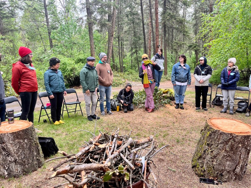 Ten humans stand in a partial circle in a forest, one speaking with their hands while wearing a fabulous jumpsuit, the others listening, rapt. A fire pit overloaded with kindling stands prominent in the foreground. 