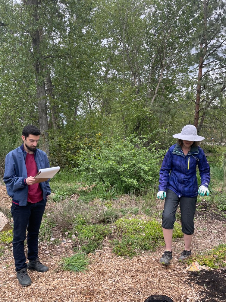 Yazdan, in a blue jacket and black pants, standing in the garden reading from a clipboard. Elana, in a floppy white sun hat and gardening gloves stands beside him looking more interested in the gardening. 