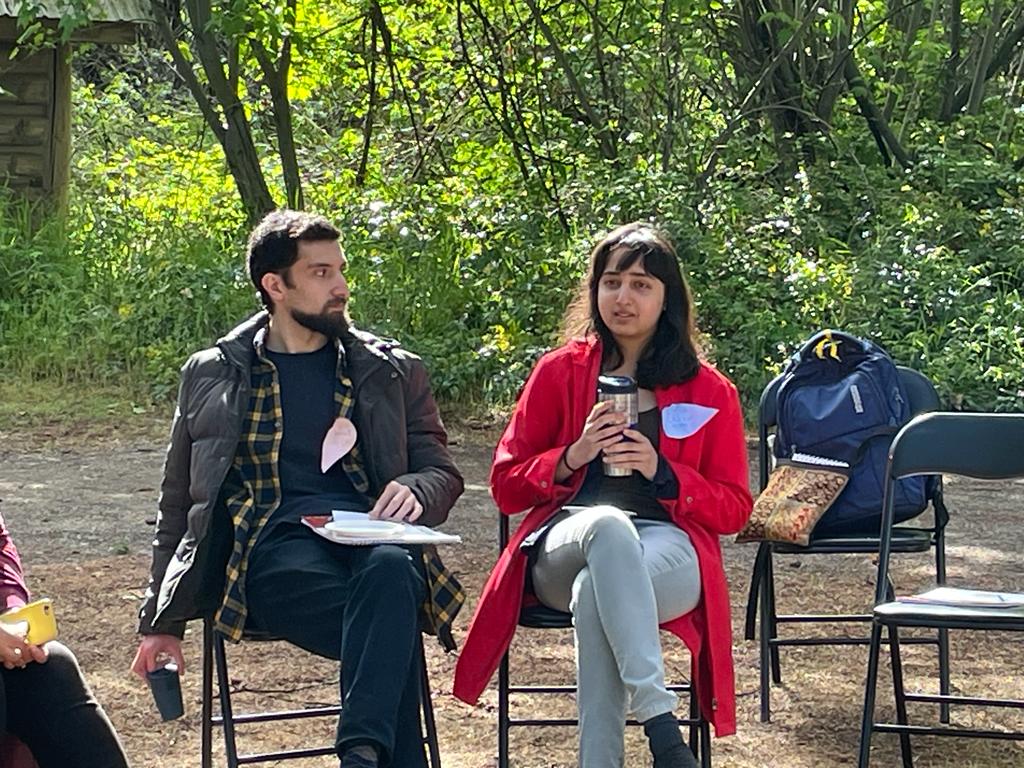 Two humans sit in folding chairs atop soil and in front of trees. They both hold beverages in reusable mugs and are dressed for chilly though well above freezing weather. One wears a red coat, the other black, both sit crossed legged and sport teardrop shaped name badges on their respective lapels. 