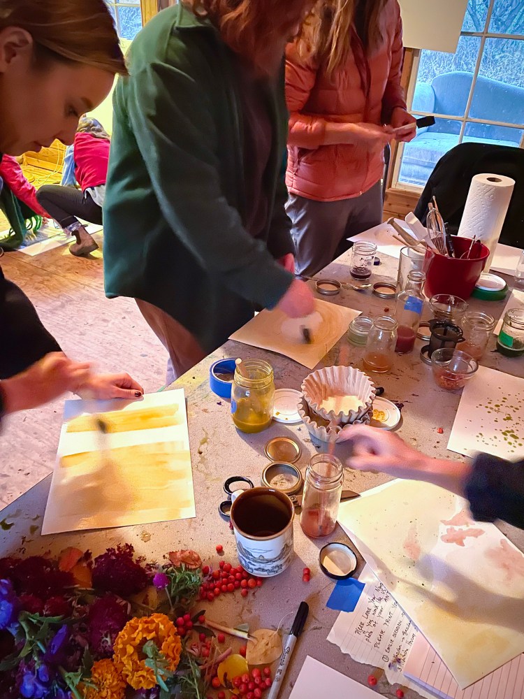 Three humans stand at a table covered with paper, pigments, brushes, glass jars, and flowers. They are painting the pigments onto white watercolour paper.