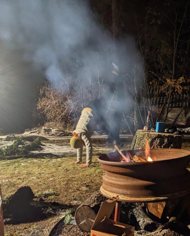 A figure in a toque bends over in a dance move, with a fire pit in the foreground. The background is the dark forest.