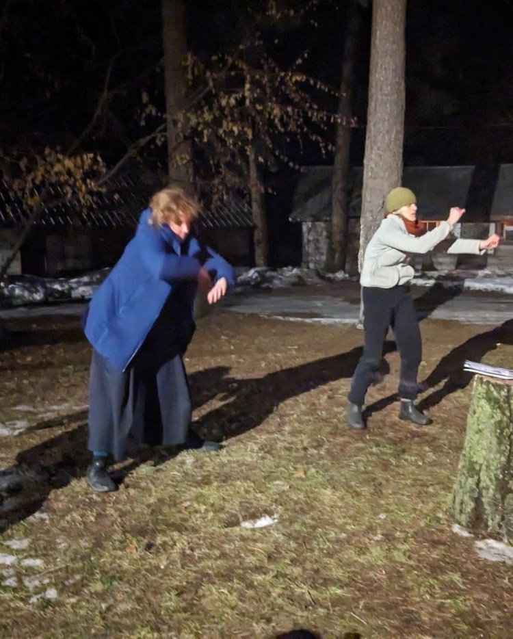 Two figures dance with their arms bent in front of them, in winter coats, in teh dark, with trees and a forest cabin in the background