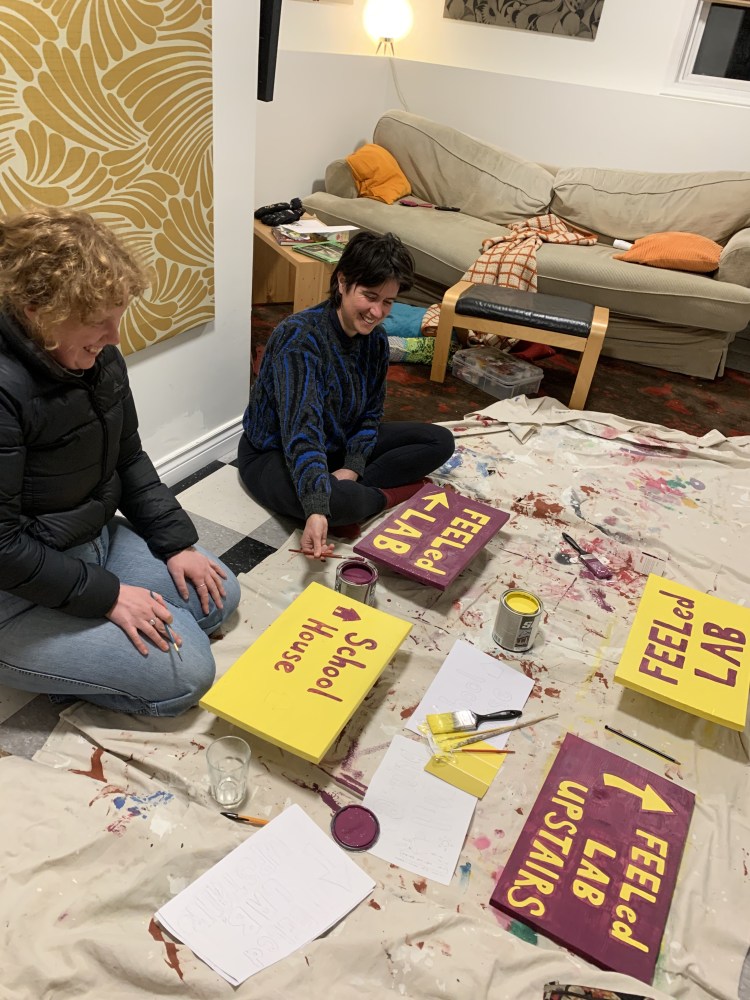 Two people in casual clothes kneel on the ground beside a paint-spattered drop sheet .They are painting purple and yellow FEELed Lab signs.
