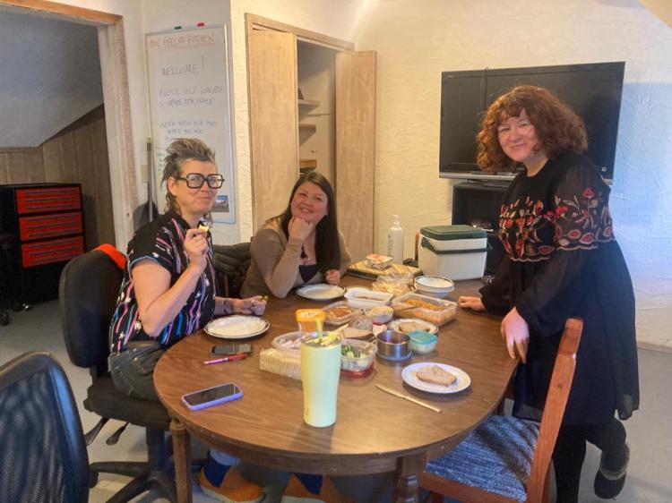 Two people sit and one person stands at a table covered in food and plates.