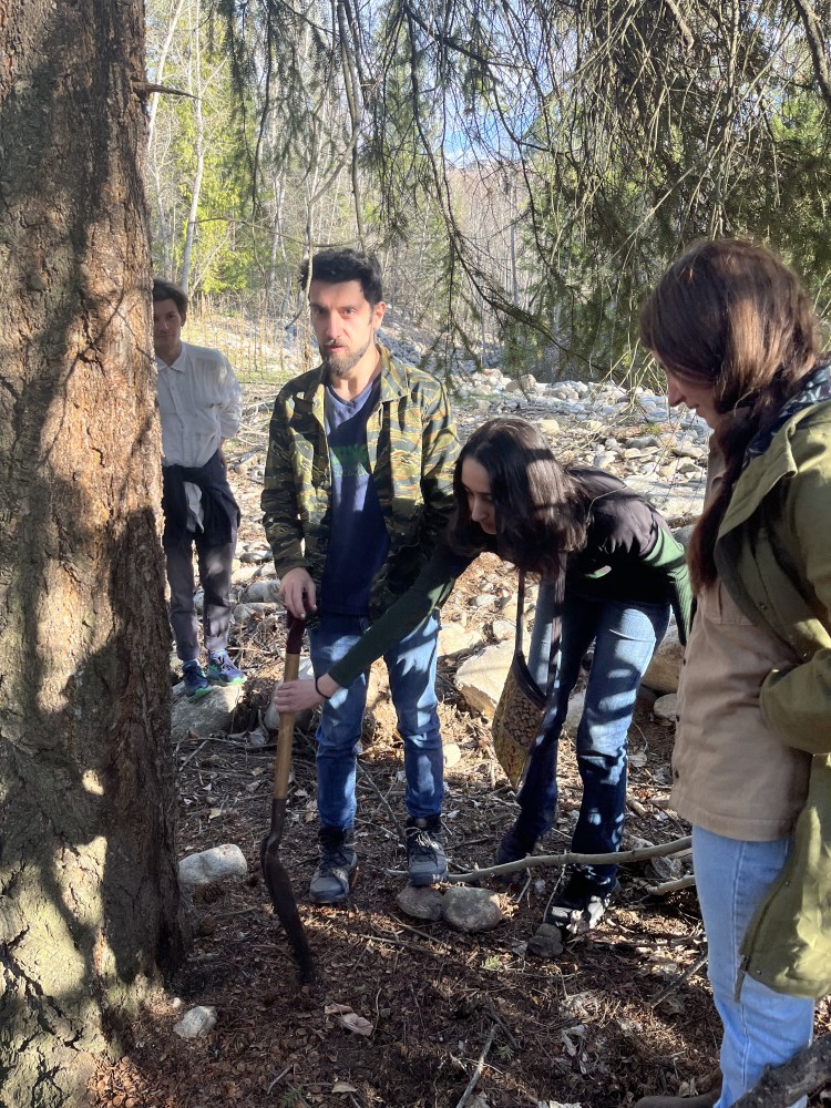 Four people stand under a tree, gathered around a shovel stuck in the ground.
