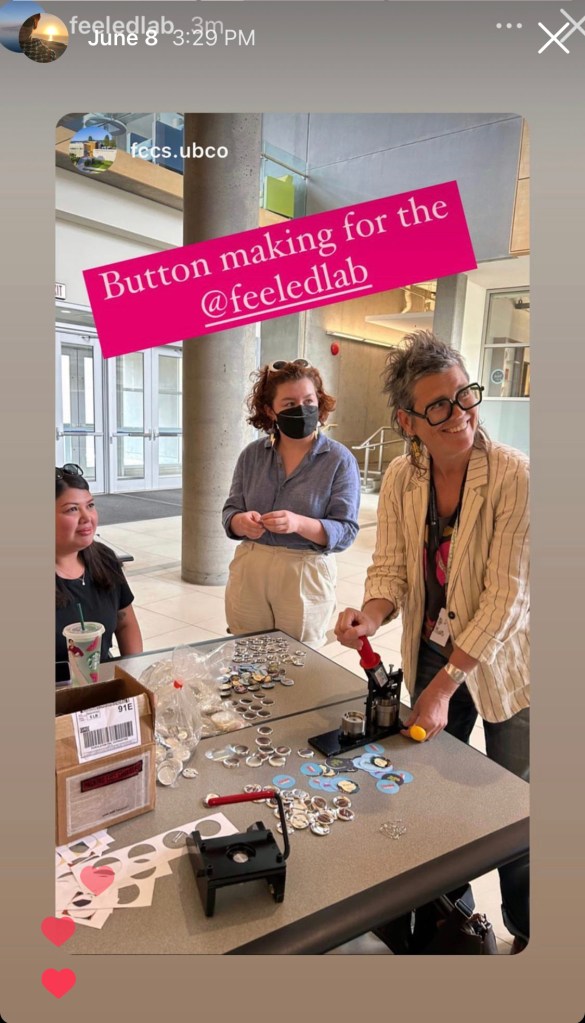 Three indivudals in the process of using a button machine at a table in an institutional-looking hallway. They all look sideways towards someone out of view.