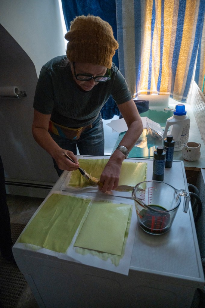 A person in a brown hat uses a paintbrush in a dark room to prepare yellow pieces of fabric and paper for cyanotype printing.