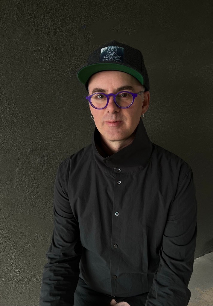 A portrait of Michael V. Smith, a white man in purple eyeglasses and a black shirt, set against a dark grey background.