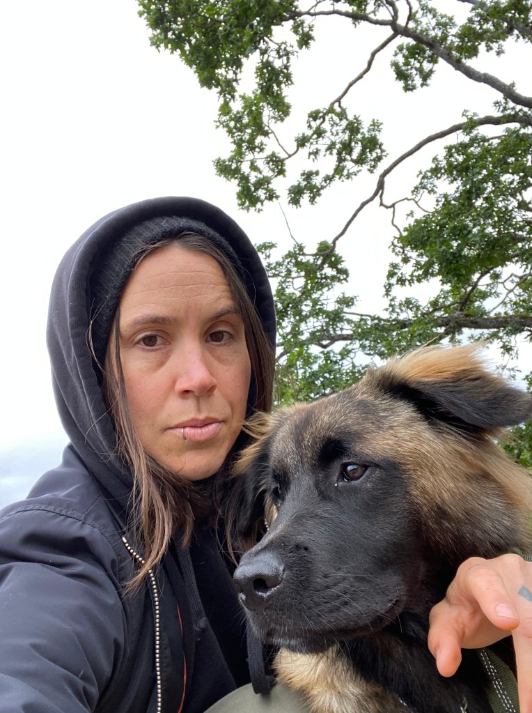 A selfie of Onyx, wearing a dark sweater and beanie, with their dog in front of some trees
