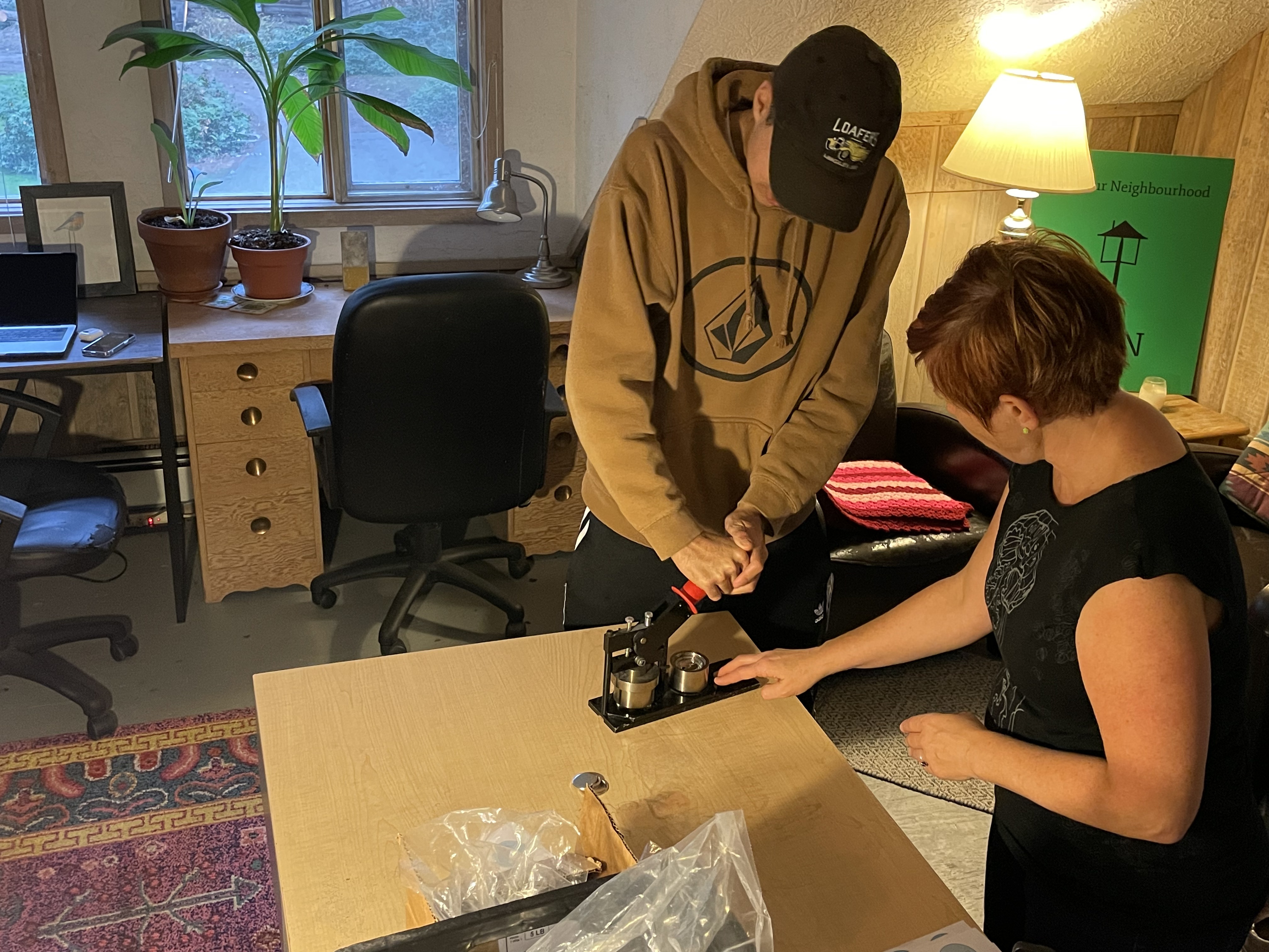 Two people stand over a table, using a button making machine!