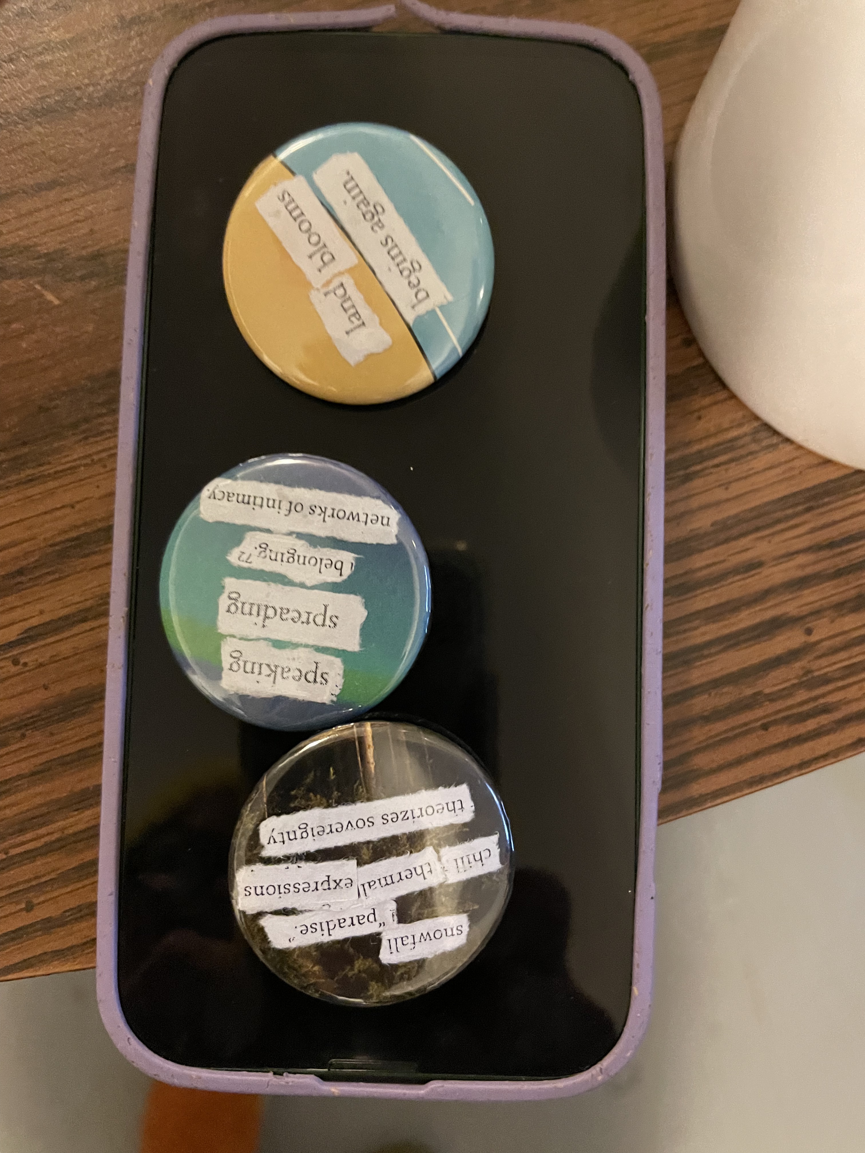 three buttons are on a mobile phone. the first reads: land blooms / begins again" ; the second: "speaking / spreading/belonging/ networks of intimacy" and the third: "snowfall / paradise/ chill thermal expressions / theorizes sovereignty"