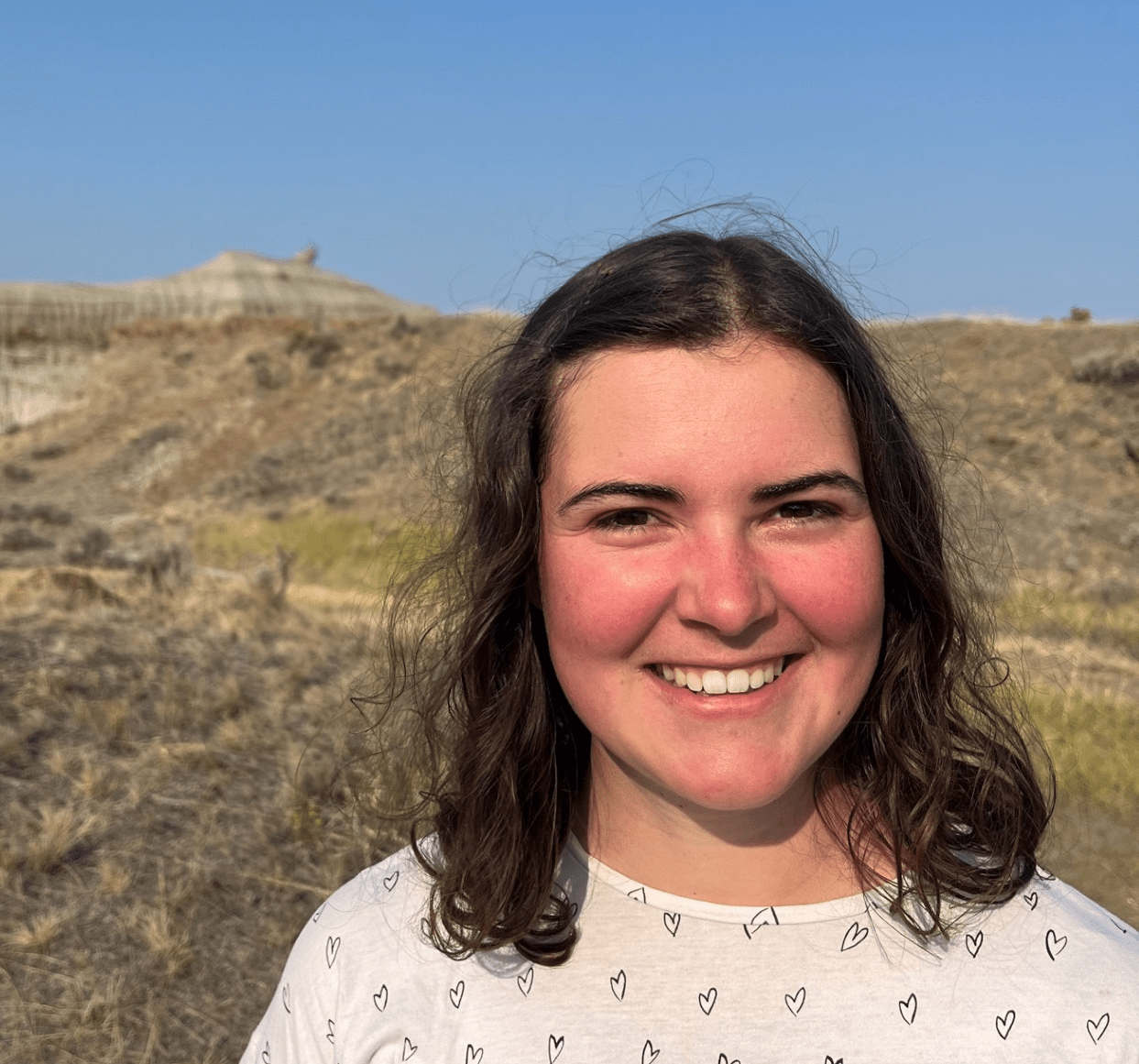A smiling white woman, rosy-cheeked with medium brown curly hair and wearing a white shirt with small hearts. Background is a desert in Southern Alberta, with brown grasses and sand formations with intersecting red rust lines.