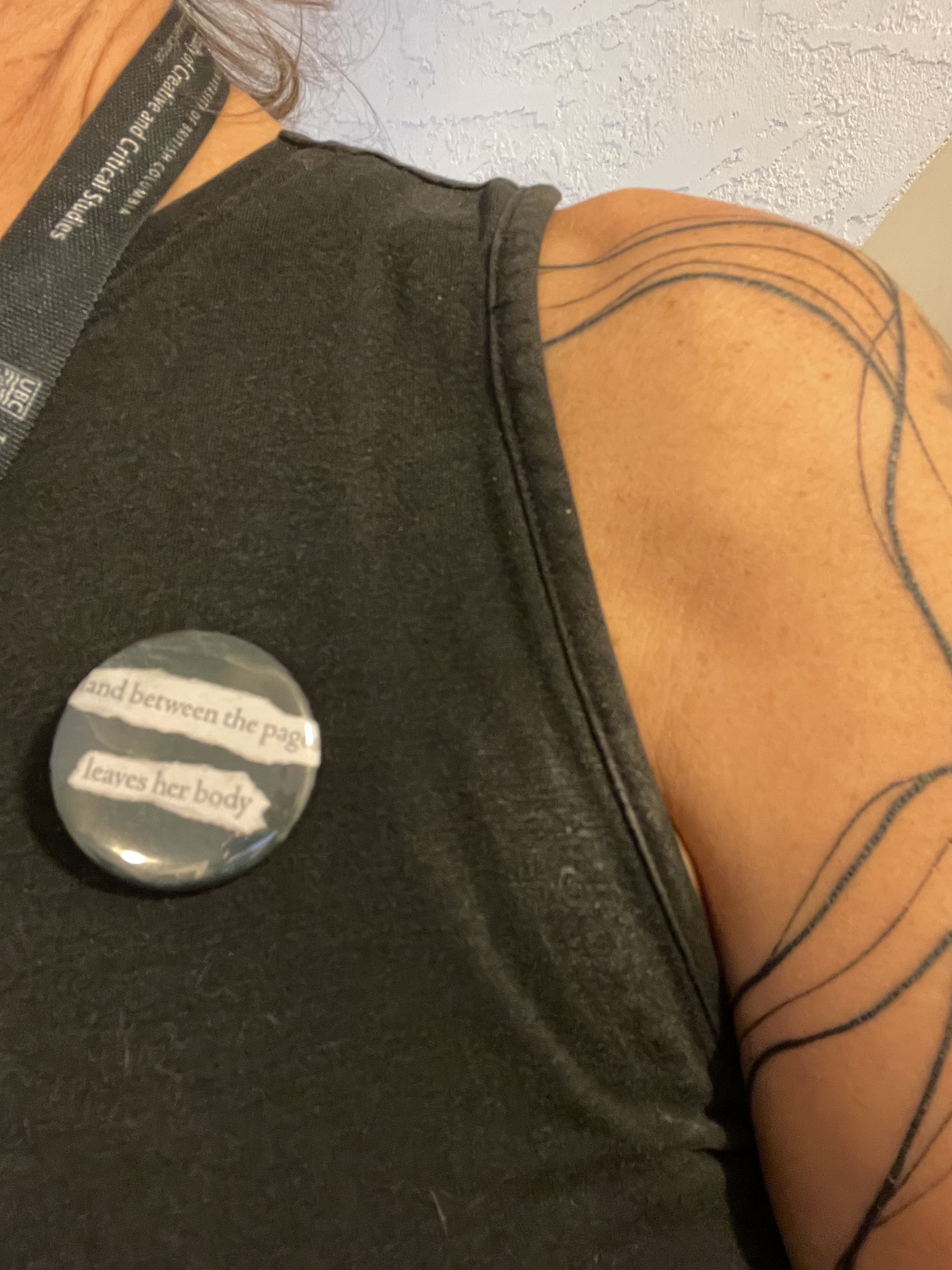 A shoulder, and a black tank top, with a button pinned to it that reads: land between the pages / leaves her body
