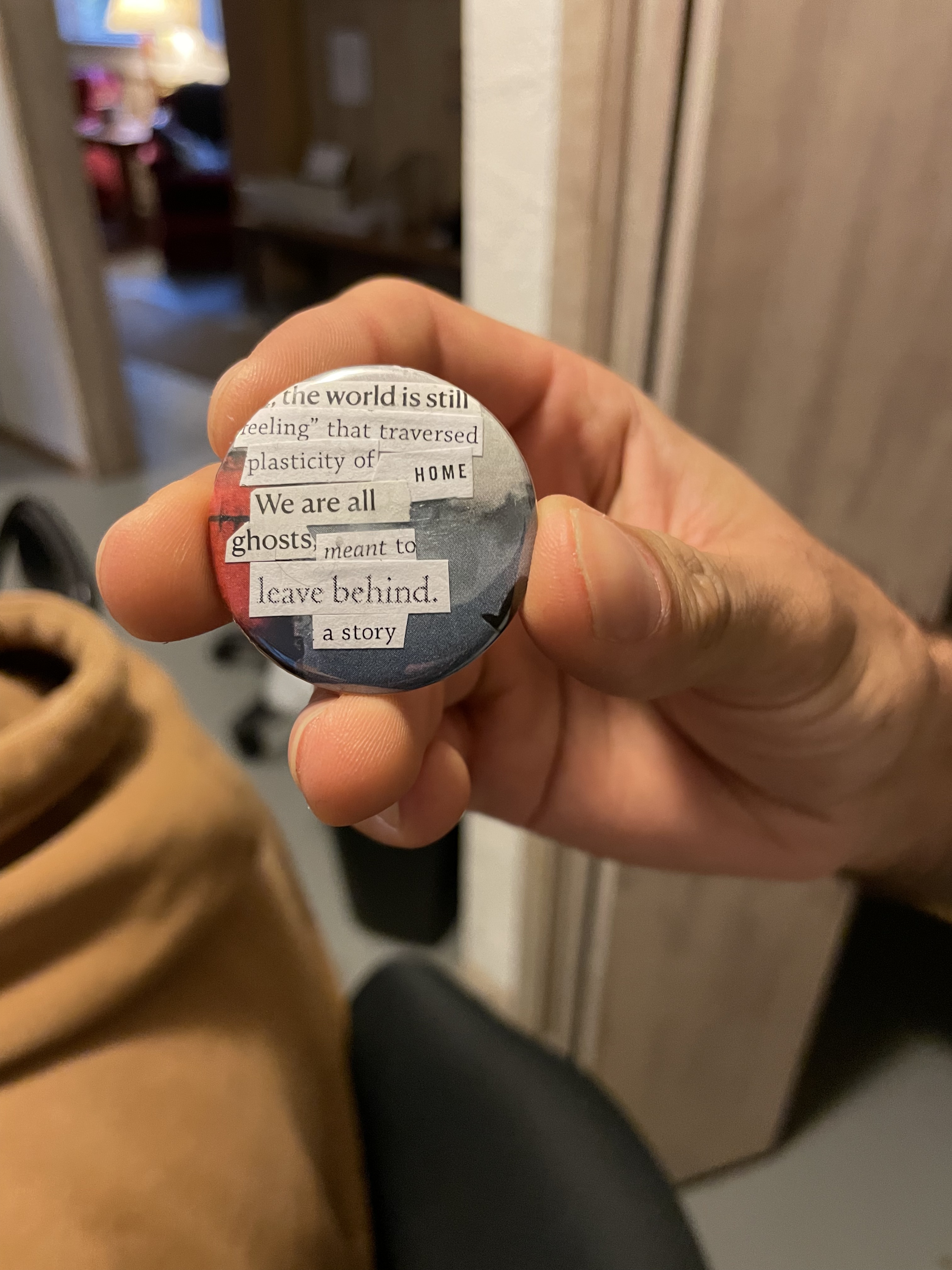 A hand holds up a button with the pastiched text "the world is stil / reeling that traversed plasticity of HOME we are all ghosts meant to leave behind a story"