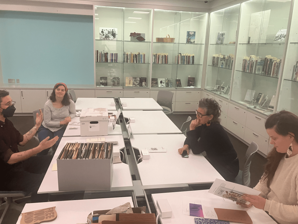 Four people sit around a large white table, with large glass display cases full of books behind them. Matt (a white man) gesticulates are he discusses a point he wants to get across as Astrida, Jenica, and Emma (three women team members) listen attentively.