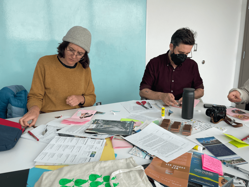Natalie wears a mustard yellow sweater and grey tuque and Matt wears a deep maroon long-sleeve shirt that is rolled up to the elbows. They both look intently at the zines in front of them, contemplating what to use for a collage from the pile of magazines in the centre of the table.