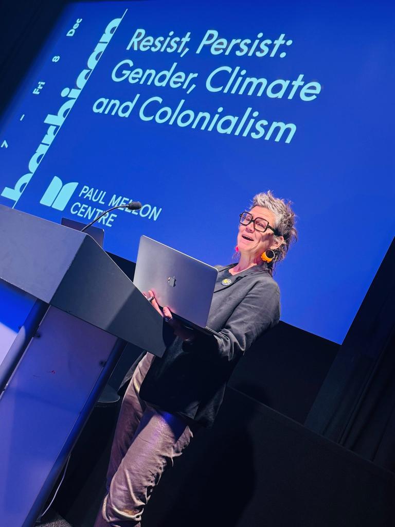 A slanted photo shows Astrida holding a laptop at a podium; behind her a big blue screen shows the text "Resist, Persist: Gender, Climate and Colonialism"
