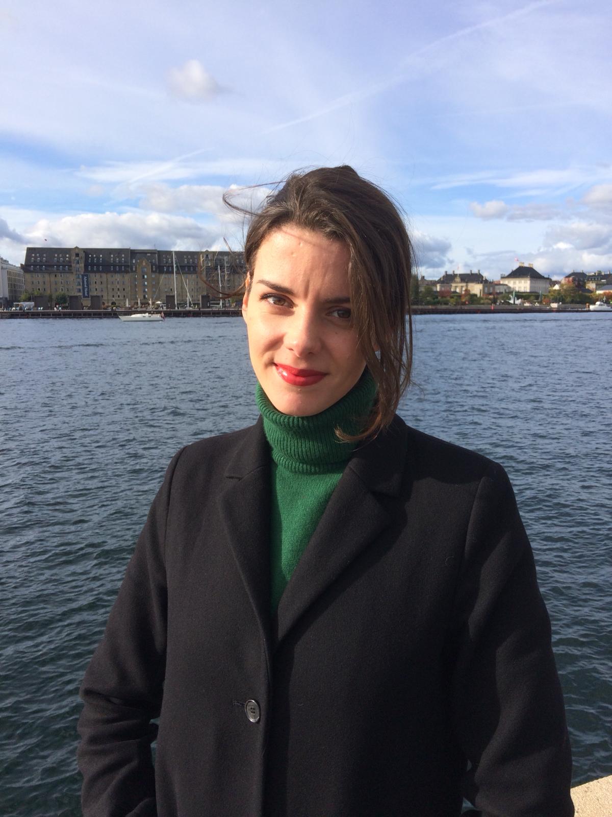 Jasmijn, a woman with brown hair and bright lipstick, softly smiling while standing in front of water bundling ina green turtle neck and dark coat.