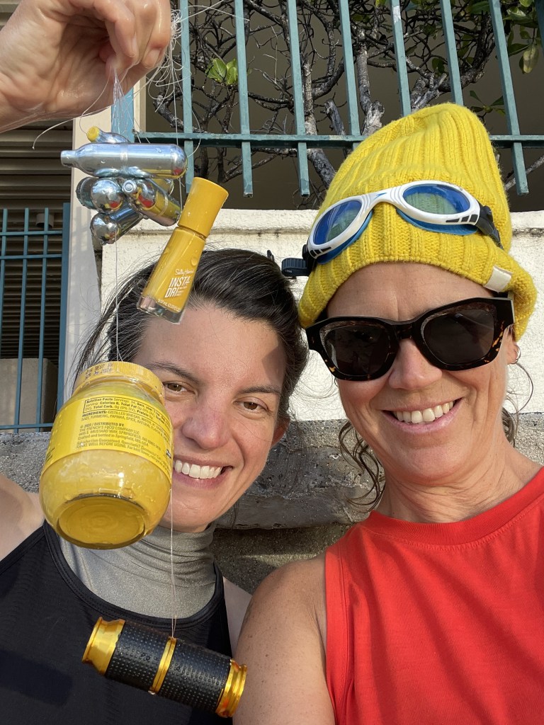 To people smile as they hold up a tangle of fishing wire holidng nail polish, nangs and a jar of mustard.