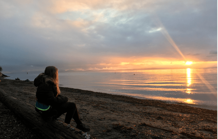A person with long hair sits in profile in a warm hoodie, on a log, looking at the sunrise across a mostly still sea.