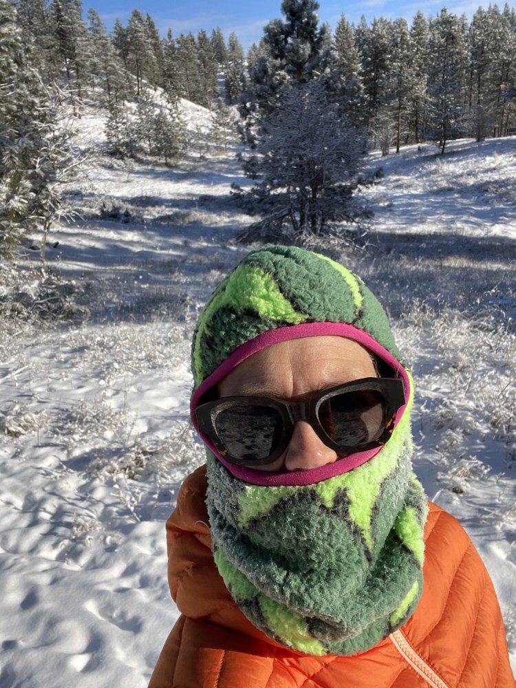 A person wearing sunglasses and a bright and fuzzy green and pink balaclava that covers their forehead, mouth and chin stands against a snowy backdrop.