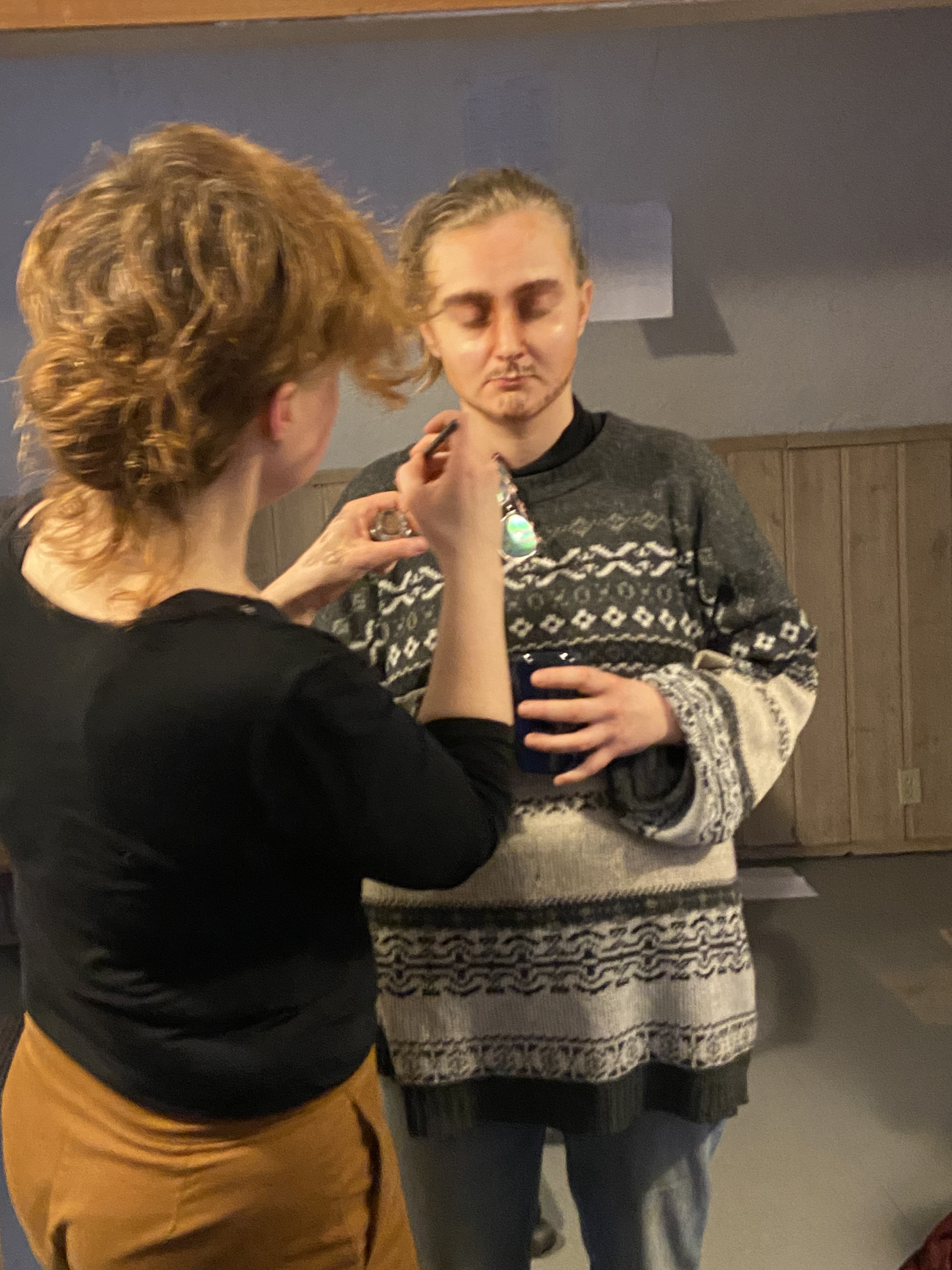 A person with auburn hair applies make-up to a person with blonde hair pulled back in a ponytail. So fa