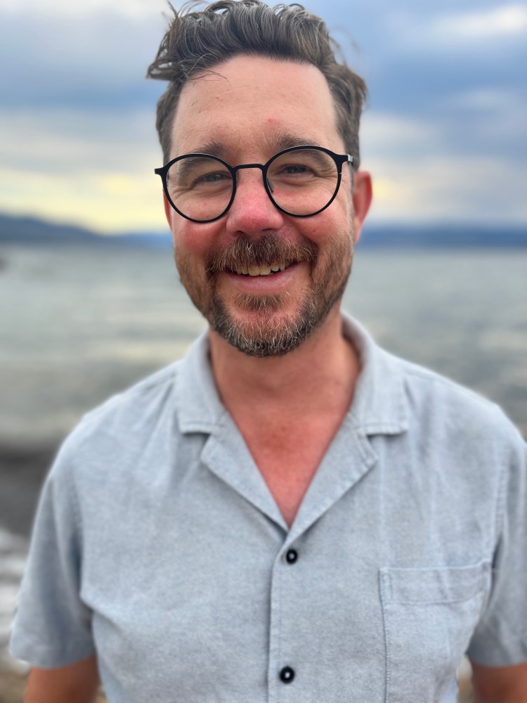 A white man with black glasses and a brown beard, smiles broadly while standing against a blurry blue background of a lake and mountains in the distance. He wears a pale blue linen shirt.