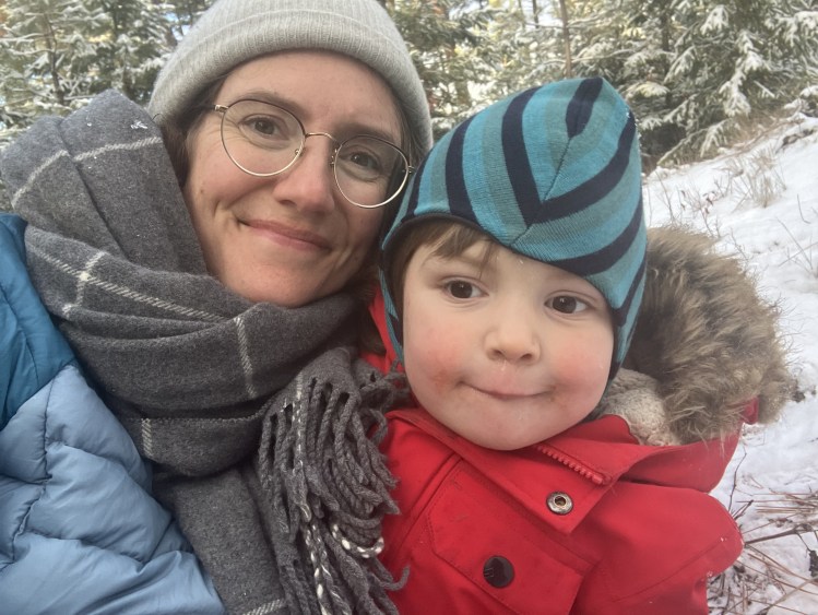 A white woman wearing glasses stands in a winter snow-covered forest hugging her child, with evergreen trees in the background. She wears a blue coat and cozy grey scarf. The child smiles cutely, wearing a bright red coat and stripy blue hat.