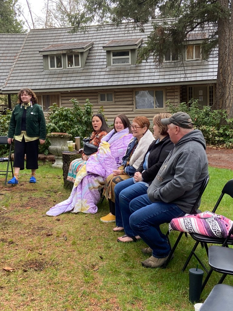 A person stands outside, with four individuals seated in chairs beside them. Someone is wrapped in a blanket and everyone looks cheerful and expectant!