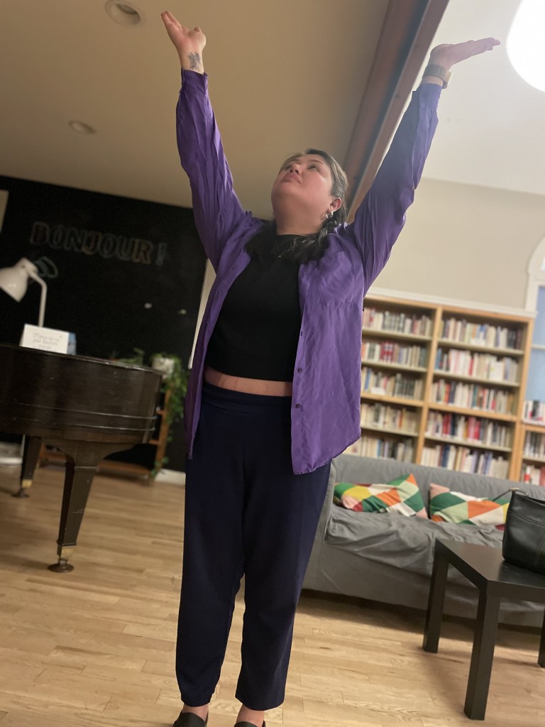 A figure dressed in black, with a purple button up shirt-coat, looks upwards with their arms extended.