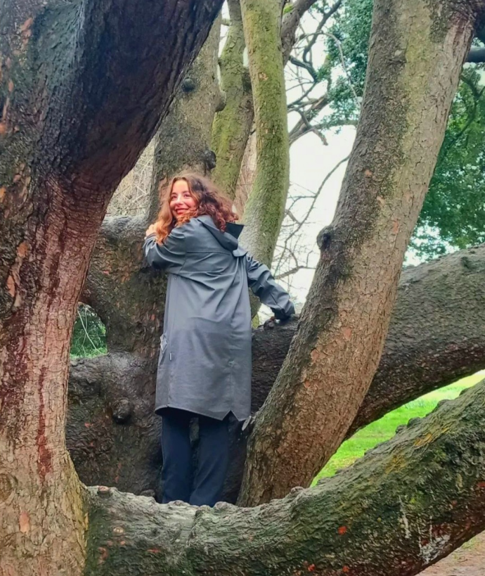 A person stands smiling in the branches of a large tree. They wear a dark raincoat and black pants, looking over their shoulder, with tree bark and foliage in the background.