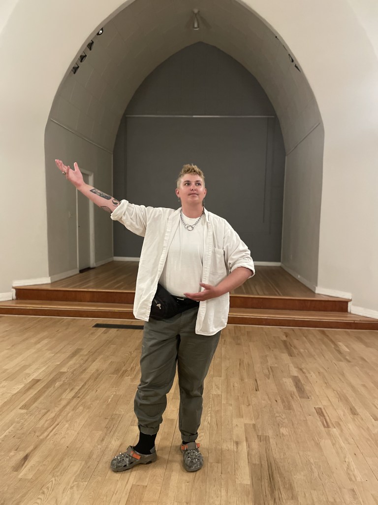 A person in khakis and a white shirt, with short hair and tattooed arms, holds one arm outstretche in a confident pose.