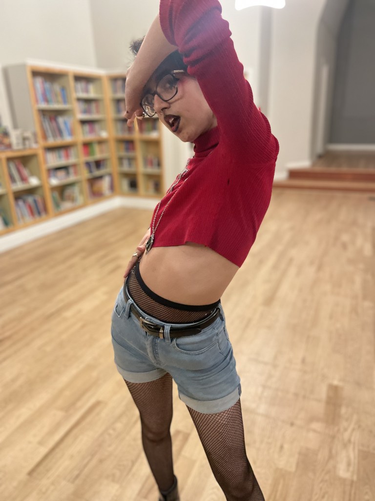A person with jean shorts over fishnet tights, with glasses and dramatic lipstick and a red crop top, stands with their hip jutted out and a bent arm over their brow.