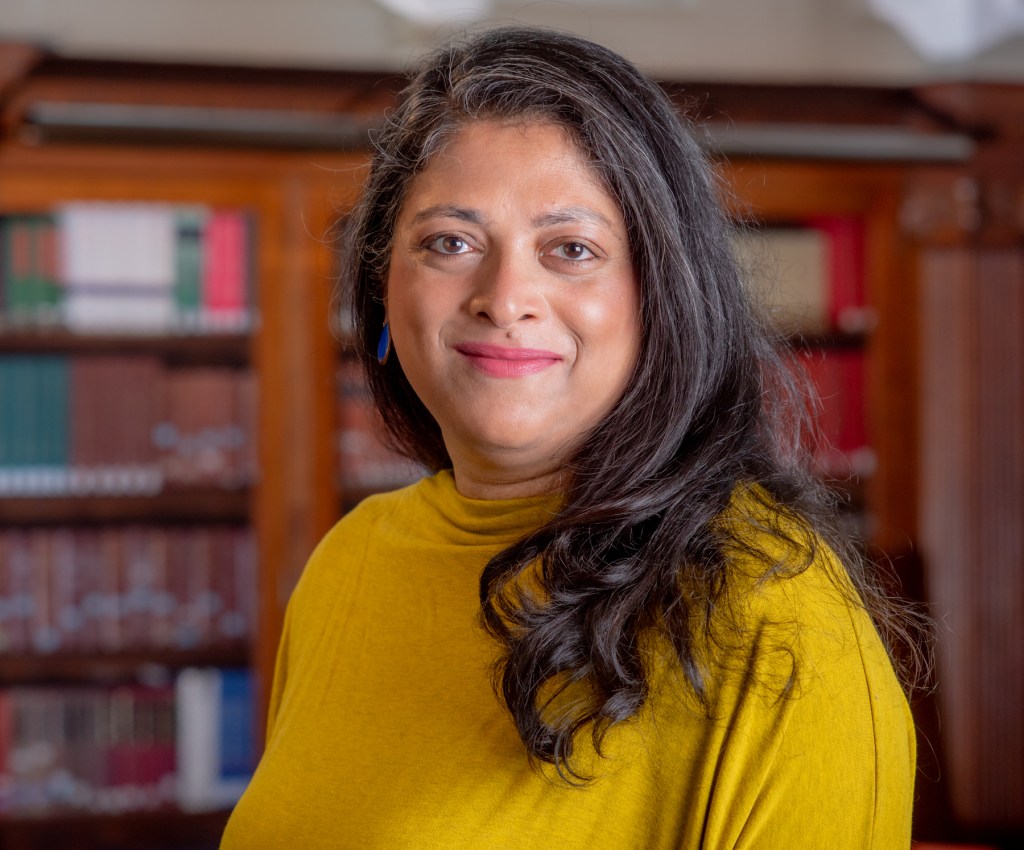 Headshot of Xan Chako wearing a yellow shirt in front of a blurred book cabinet.
