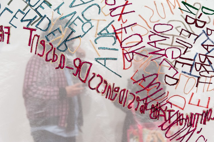 two people talking behind a semi-transparent screen, on the screen are large red, peach and teal coloured letters