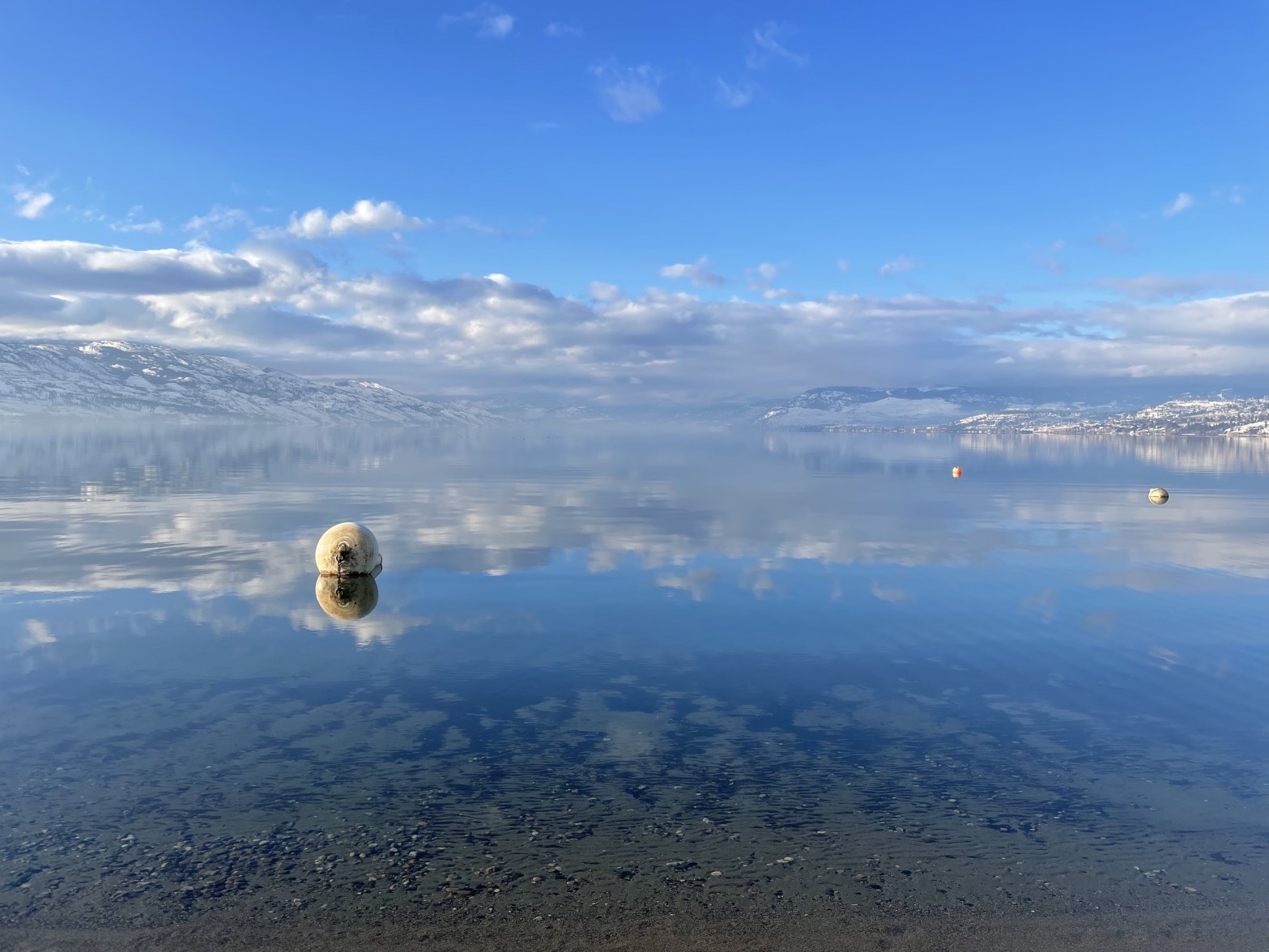 A large body of water with snow-capped mountains in the background, the clouds, sky and mountains are reflected in the lake giving it a dreamy quality