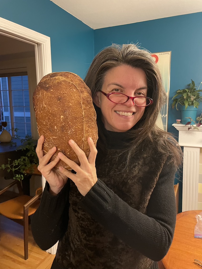 A dark haired woman with glasses holds up a loaf of bread close to her face