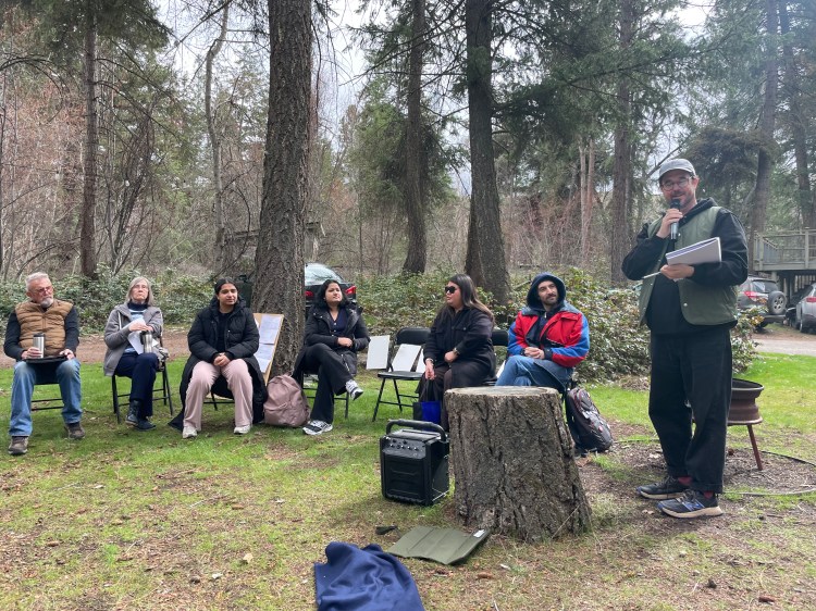A group of people gather in a wooded clearing. One man with glasses and a ball cap reads is standing, reading from his notes.