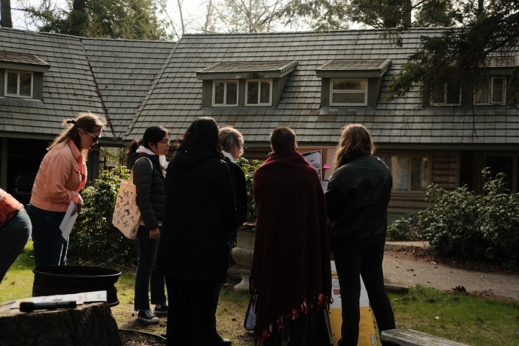 A half-circle of people gather around a bulletin board with stories pinned to it. They silently read. A couple Oregon grape bushes observe on both sides of them. A house with a green-roof sits in the background.