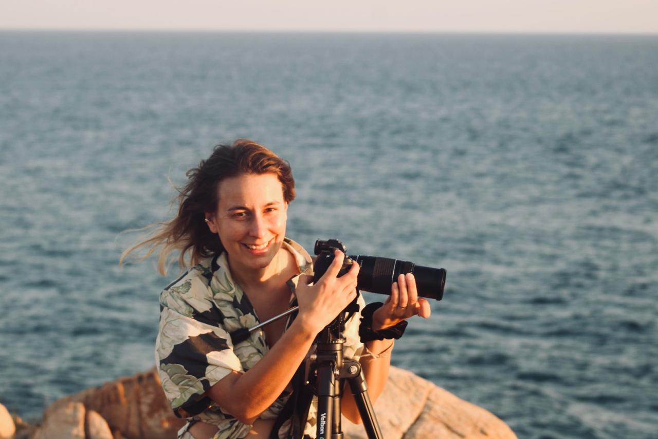 headshot of Clara in front of a large body of water holding a camera and smiling