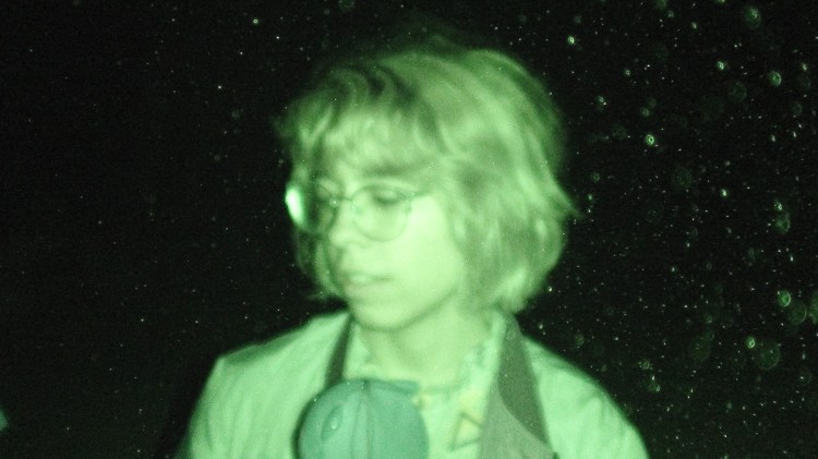 An eerie photo at night of a green-tinged person with classes and light chin-length hair, looking down and off to the side