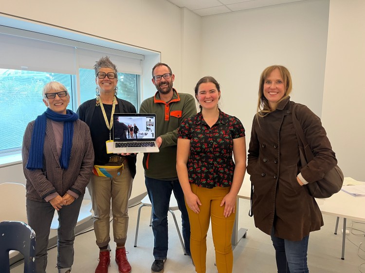 5 people stand in a line smiling, with a laptop held up too 